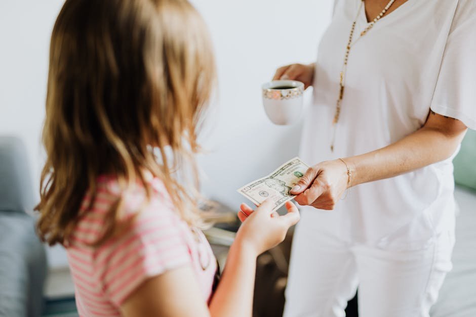 A woman hands a dollar bill to her daughter while holding a coffee cup, indoors.